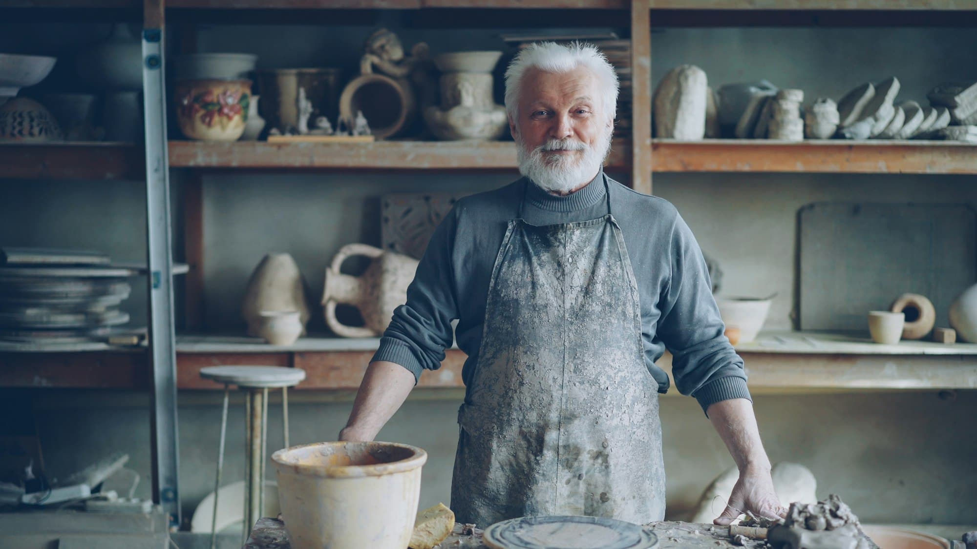 A potter in a clay-streaked apron stands proudly in his workshop.