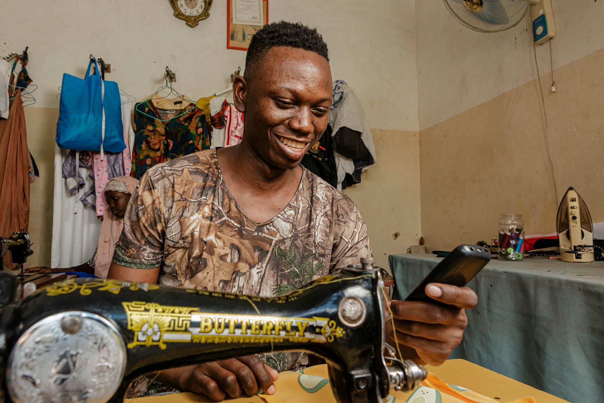 A tailor smiles at his phone while working at his sewing machine.